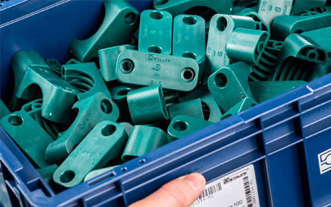 Blaue Industriekiste gefüllt mit vielen grünen Kunststoff-Rohrschellen und Befestigungselementen der Marke Stauff.