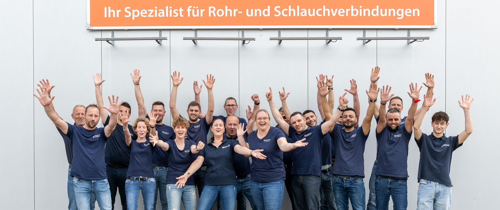 Mitarbeiter des Schellen-Shops stehen unter einem Banner mit dem Slogan "Ihr Spezialist für Rohr- und Schlauchverbindung"